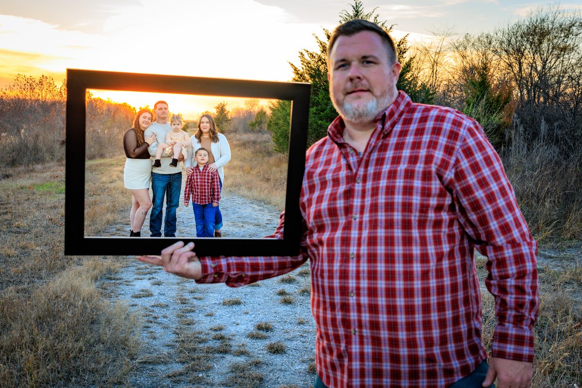 Man in a plaid shirt holding an empty picture frame that frames his family standing behind him on a trail at sunset