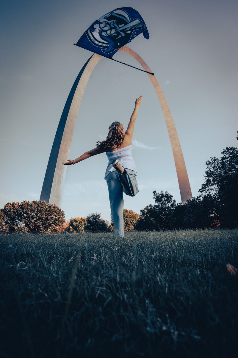 High school senior seen from behind, kicking up one leg and reaching toward a school flag tossed high under the Gateway Arch